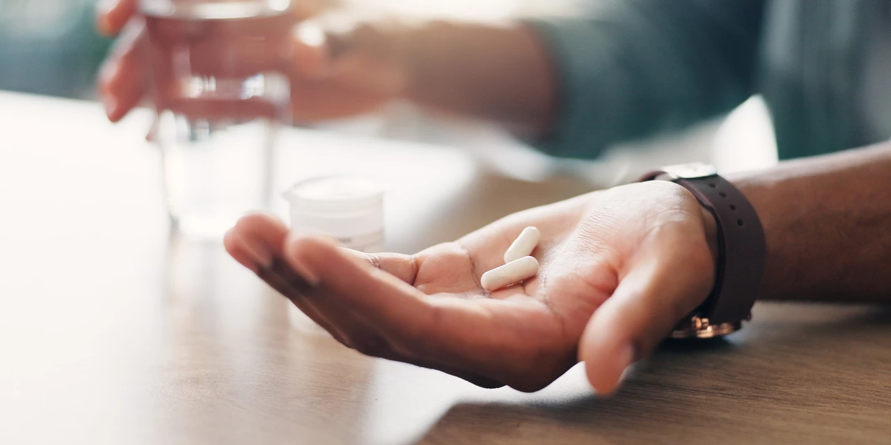 A person holds prescription drugs in their hand beside a glass of water, reflecting daily medication use and possible dependence on prescription drugs.