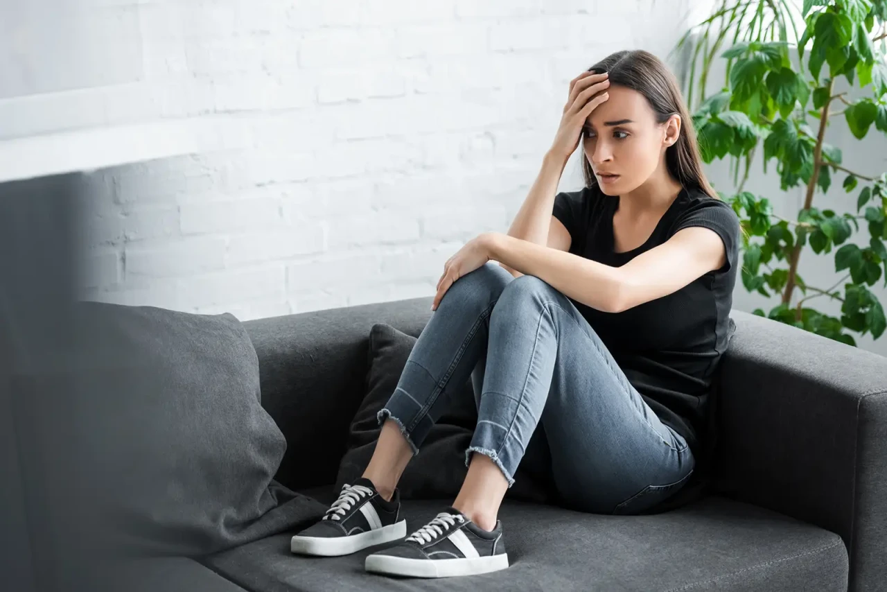  A worried woman sitting on a grey couch against a white background, with a plant behind her.