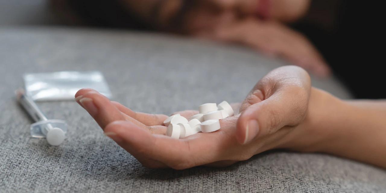 A person holds several white medicine tablets in their hand beside a syringe, illustrating behaviours linked to medicine addiction.