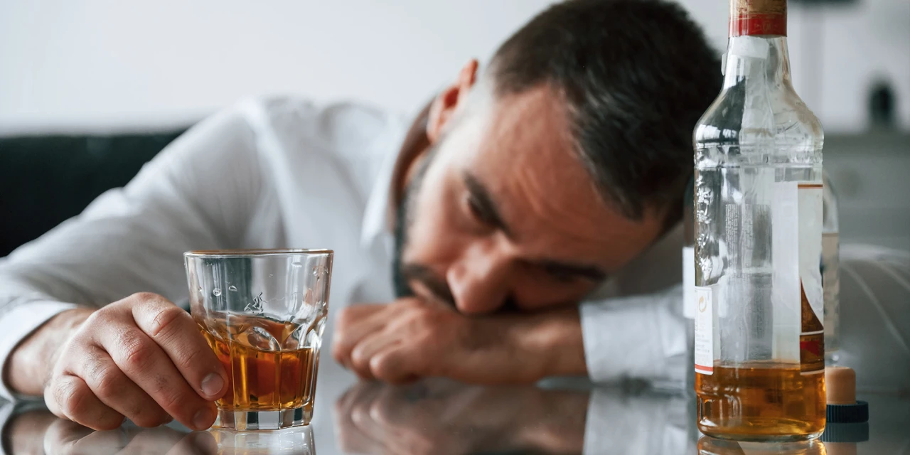 Een man ligt uitgeput naast een fles en een glas alcohol, wat de fysieke en emotionele gevolgen van alcoholverslaving in het dagelijks leven illustreert.