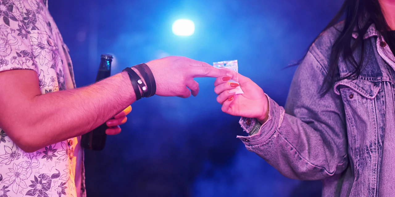 Two people exchange a small bag of drugs in a nightclub setting, reflecting the social environments often associated with ecstasy addiction.
