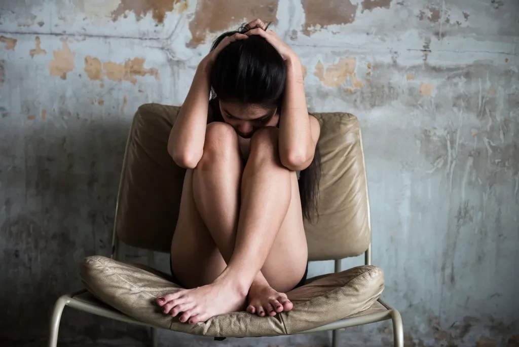 woman-hugging-herself-on-chair A woman hugging herself and holding her head, sitting on a chair with her legs folded against an industrial wall.