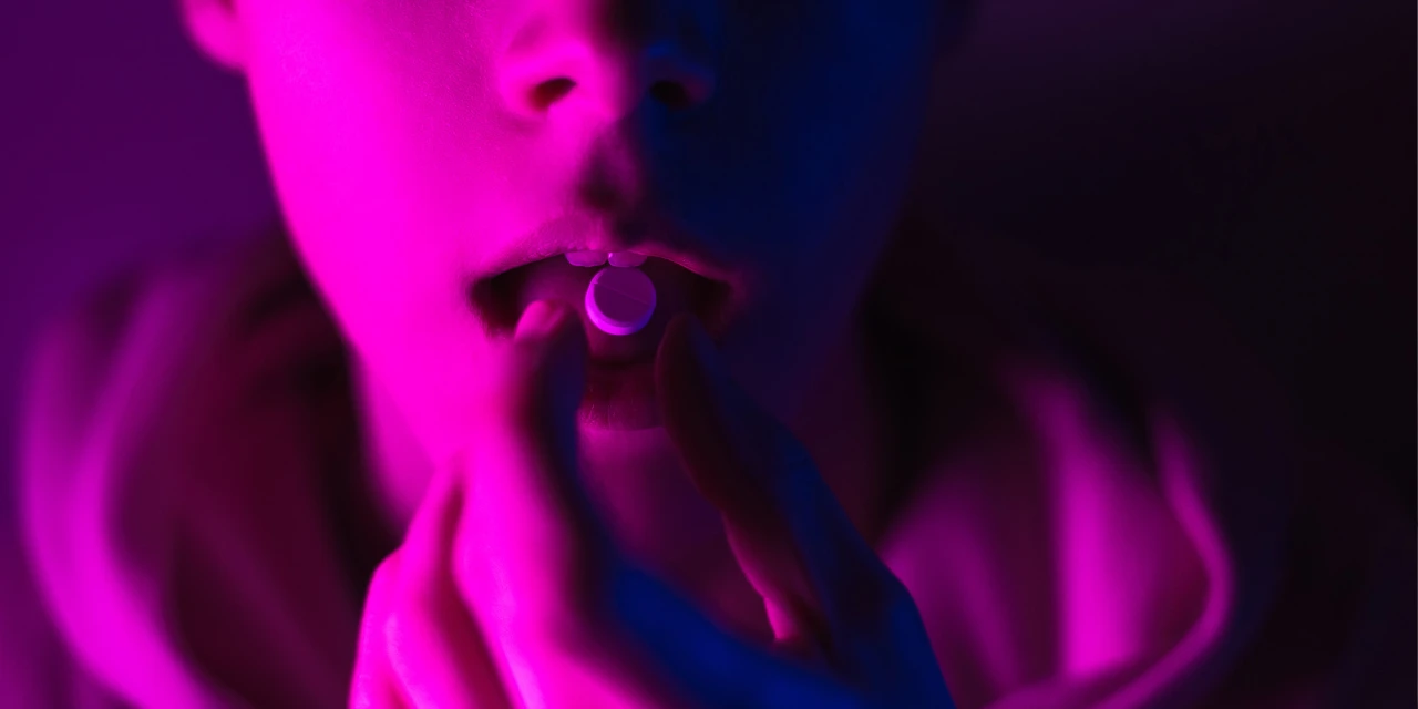 A person places a pill on their tongue under neon lighting, showing recreational drug use associated with LSD addiction.