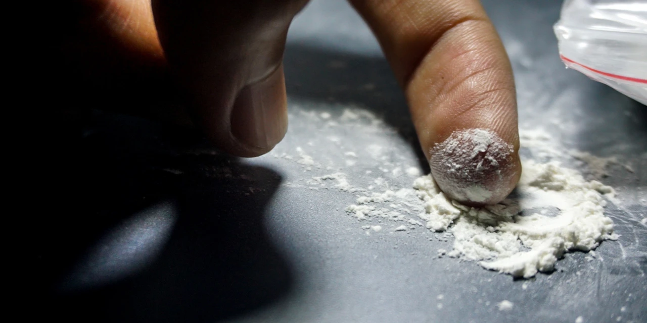 Fingertip pressed into white ketamine powder on a dark table, small plastic bag beside it.