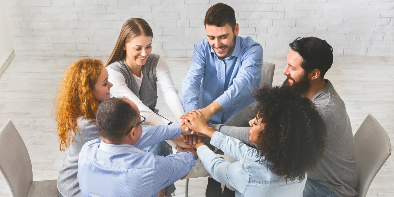 An overhead view of a group of people sitting in a circle, placing their hands together in the center.