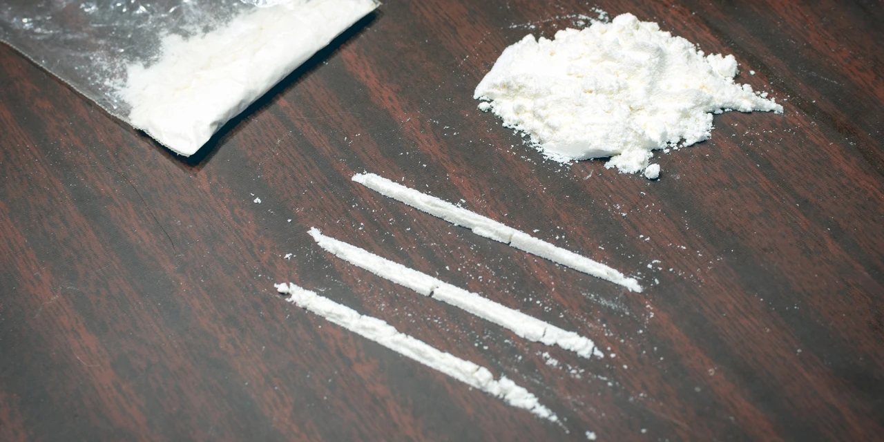 Drie lijnen wit poeder op tafel naast zakje en hoopje verwijzen naar cocaïneverslaving.