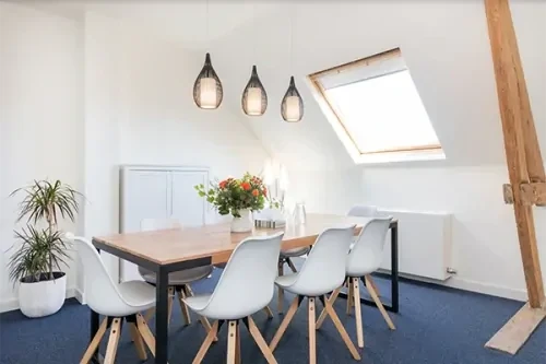 Behandelkamer met witte muren, een blauw tapijt, een houten tafel en witte stoelen en verschillende planten.