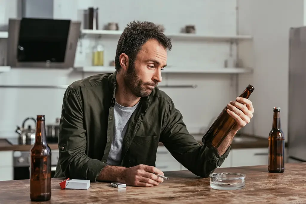 mand-looking-at-bottle A man sits at a kitchen table surrounded by empty bottles. He gazes doubtfully at a bottle in his hand.