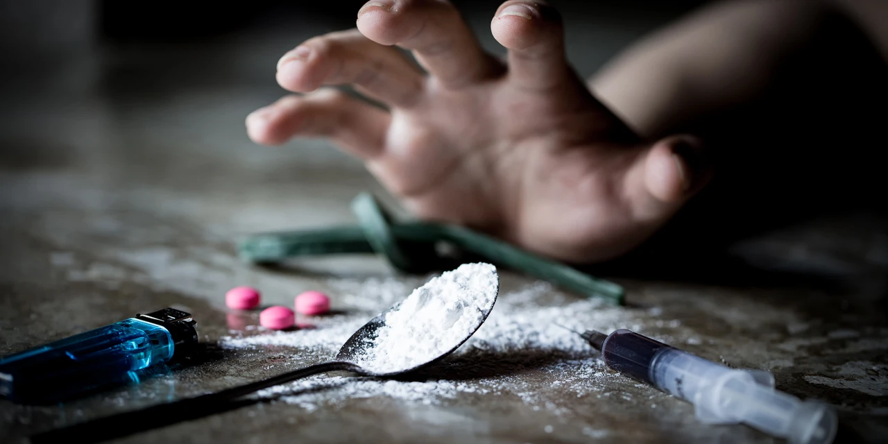 A hand reaches toward white powder, pills and a syringe on the floor, illustrating the dangers and physical risks linked to amphetamine addiction.