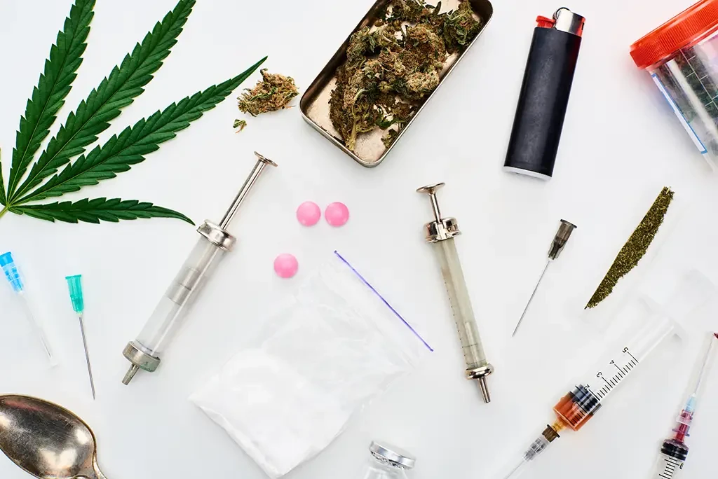 Different types of drugs laid out on a white table, including a cannabis leaf, syringes, pills, a lighter, and a bag of powder.