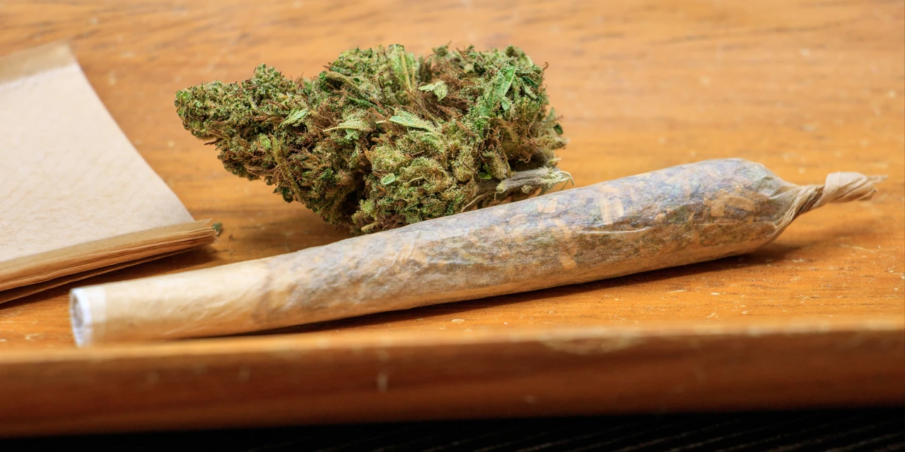 A rolled marijuana joint lies beside cannabis buds on a wooden table, representing substance use linked to marijuana addiction.