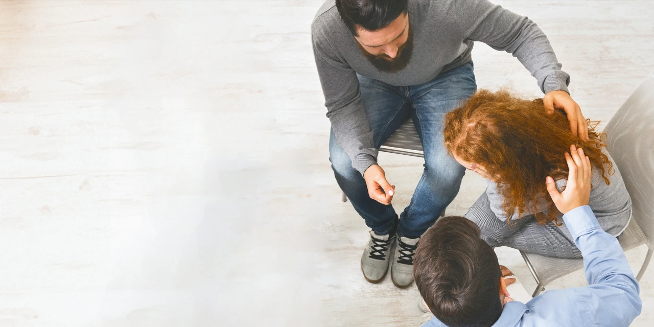A high-angle shot showing two men offering comfort to a woman who is leaning forward during a difficult moment.