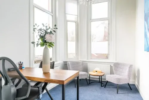 Behandelkamer met bureaustoel, houten bureau en twee fauteuils tegen hoge ramen en witte muren.