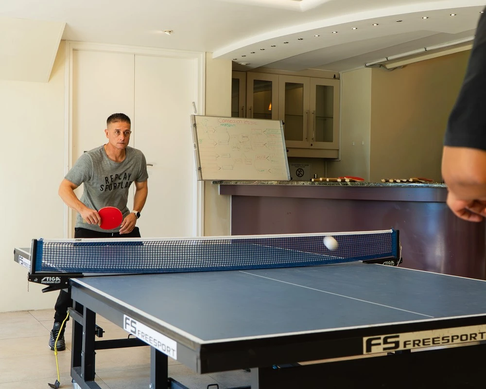 Two people are playing table tennis in a bright recreation room.