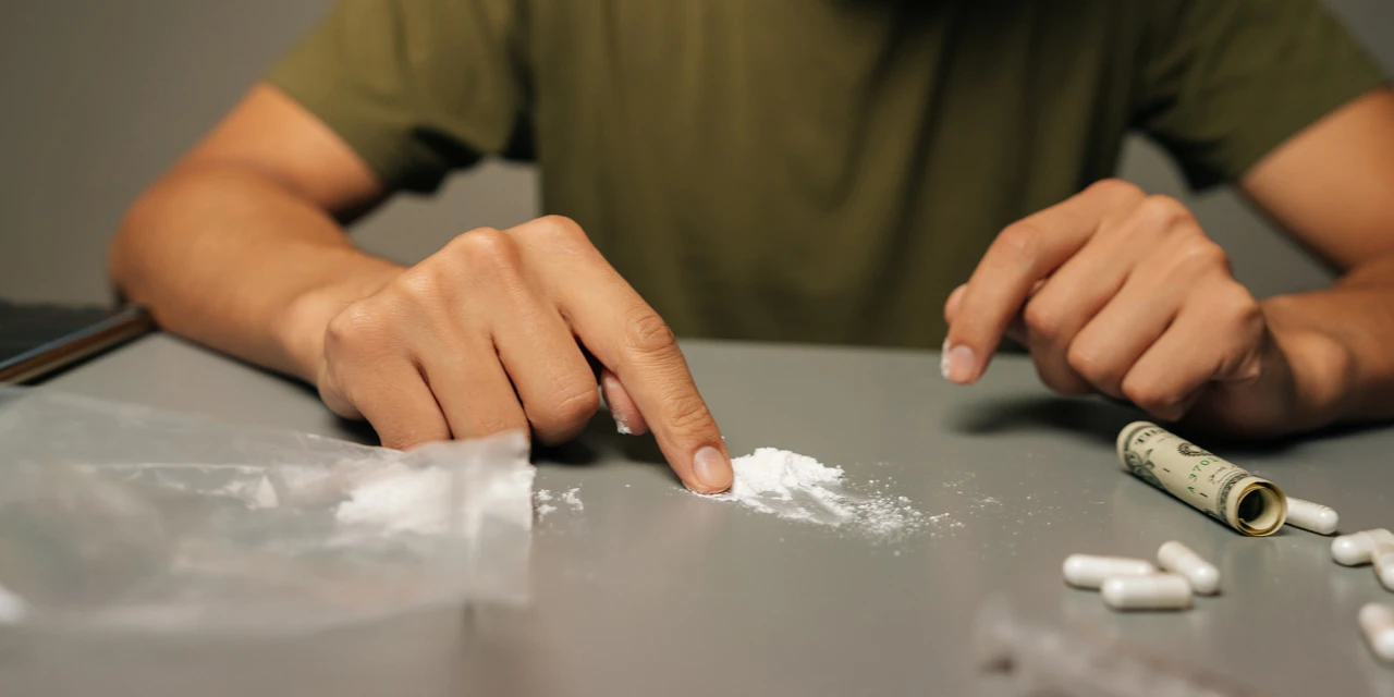 Person using cocaine on a table with white powder and pills nearby.
