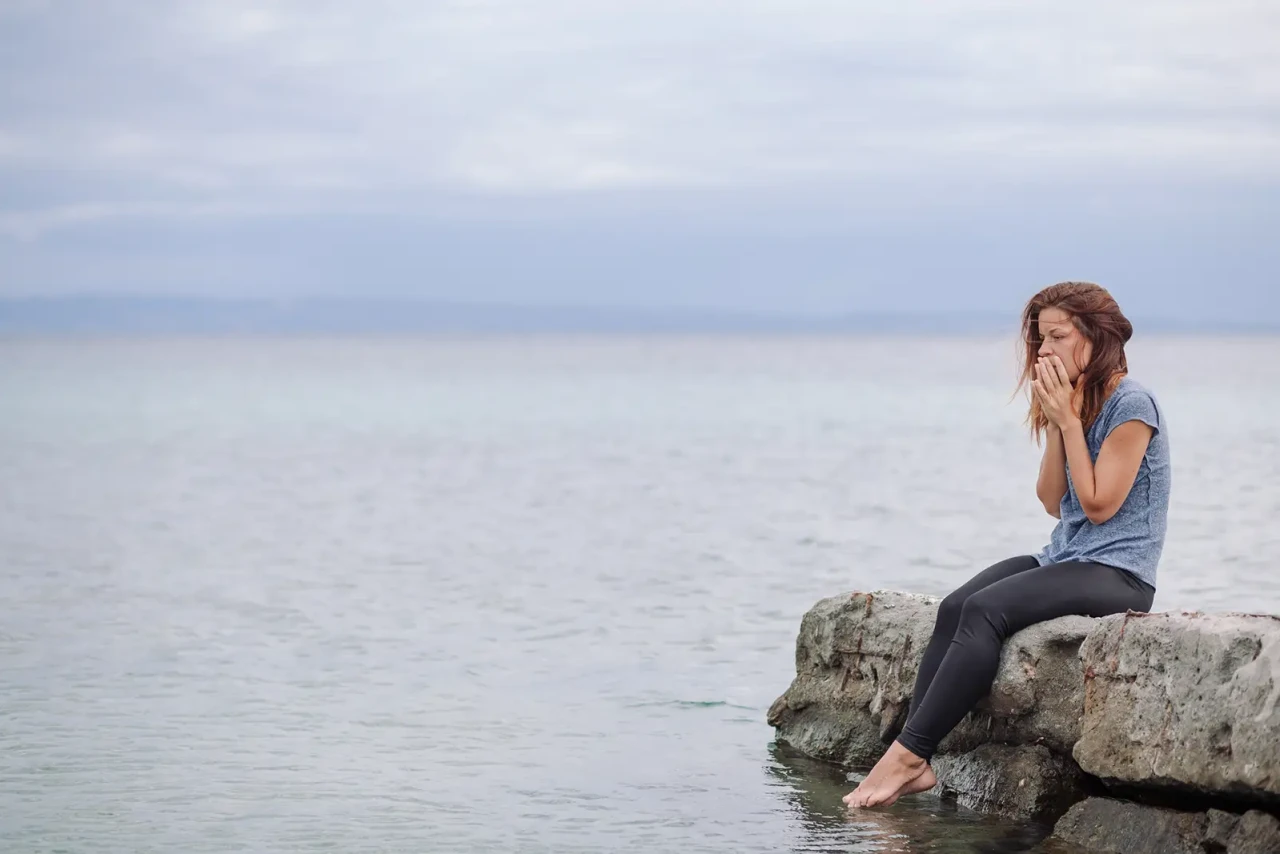 A woman sitting on rocks in the ocean, with her hands in front of her mouth.