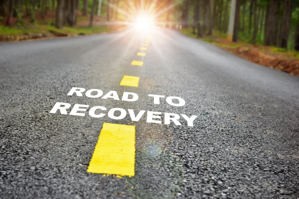 A road with yellow stripes and the text 'road to recovery', with the sun and trees in the background.