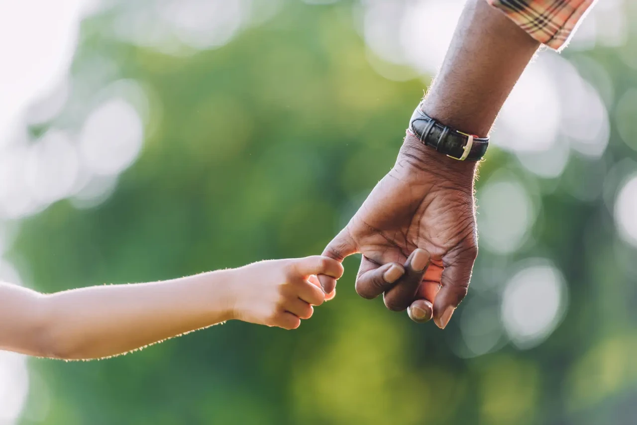 A child's hand holding a man's hand by the pinkie, with green foliage in the background.